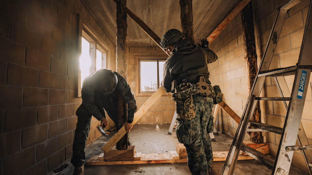 Twee militairen in een onafgewerkte ruimte: één in gevechtsuitrusting staat recht, de ander bukt om houten balken te plaatsen bij een raam.