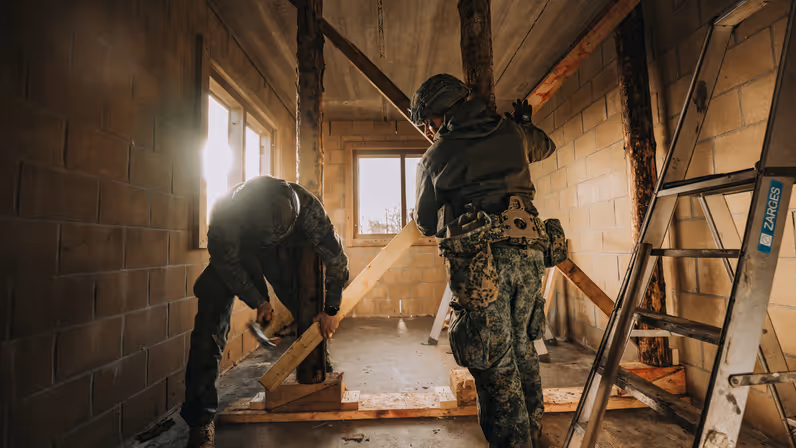 Twee militairen in een onafgewerkte ruimte: één in gevechtsuitrusting staat recht, de ander bukt om houten balken te plaatsen bij een raam.