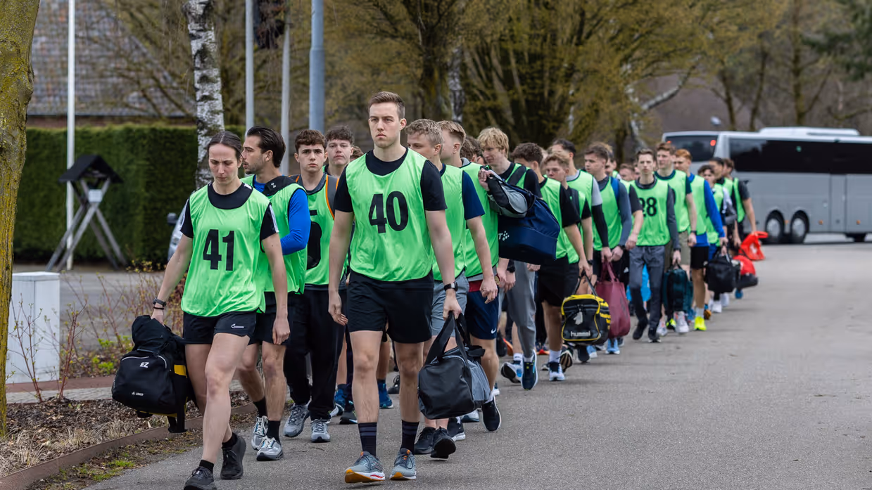 Een groep jongeren in felgroene hesjes met nummers staat in het gelid. Een militair in uniform geeft instructies op een parkeerterrein met bomen en voertuigen.