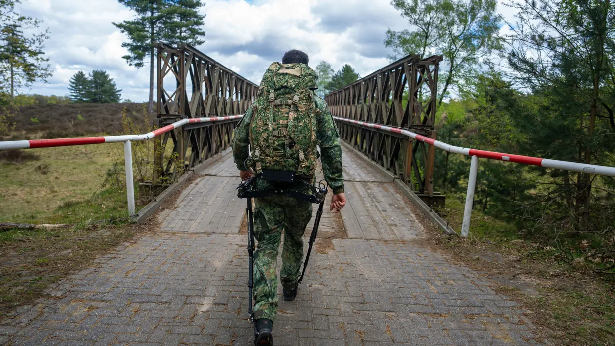 Links: een soldaat in camouflage met rugzak loopt over een houten brug met rode-witte afzetting in een bosrijk gebied.    Rechts: een soldaat in camouflage met rugzak en stokken loopt over een weg met dennenbomen en een bewolkte lucht. 
