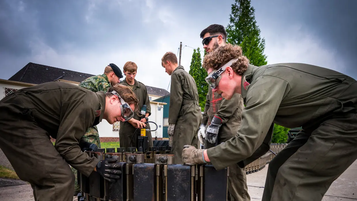Soldaten in uniform werken met zware metalen onderdelen bij een groene militaire vrachtwagen, omringd door gereedschap en uitrusting.