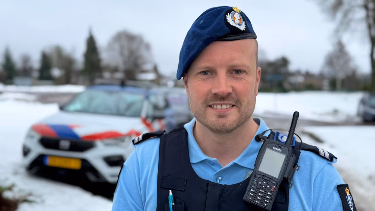 Portret van wachtmeester van de Marechaussee met voertuig op de achtergrond.