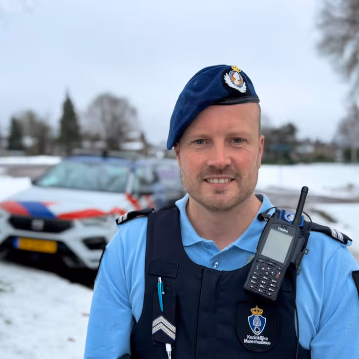 Portret van wachtmeester van de Marechaussee met voertuig op de achtergrond.