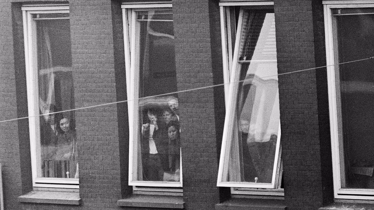 Alttekst: 3 ramen: in het middelste raam staan 2 mensen achter het glas, het rechterraam is kapot en de gordijnen wapperen naar buiten. 