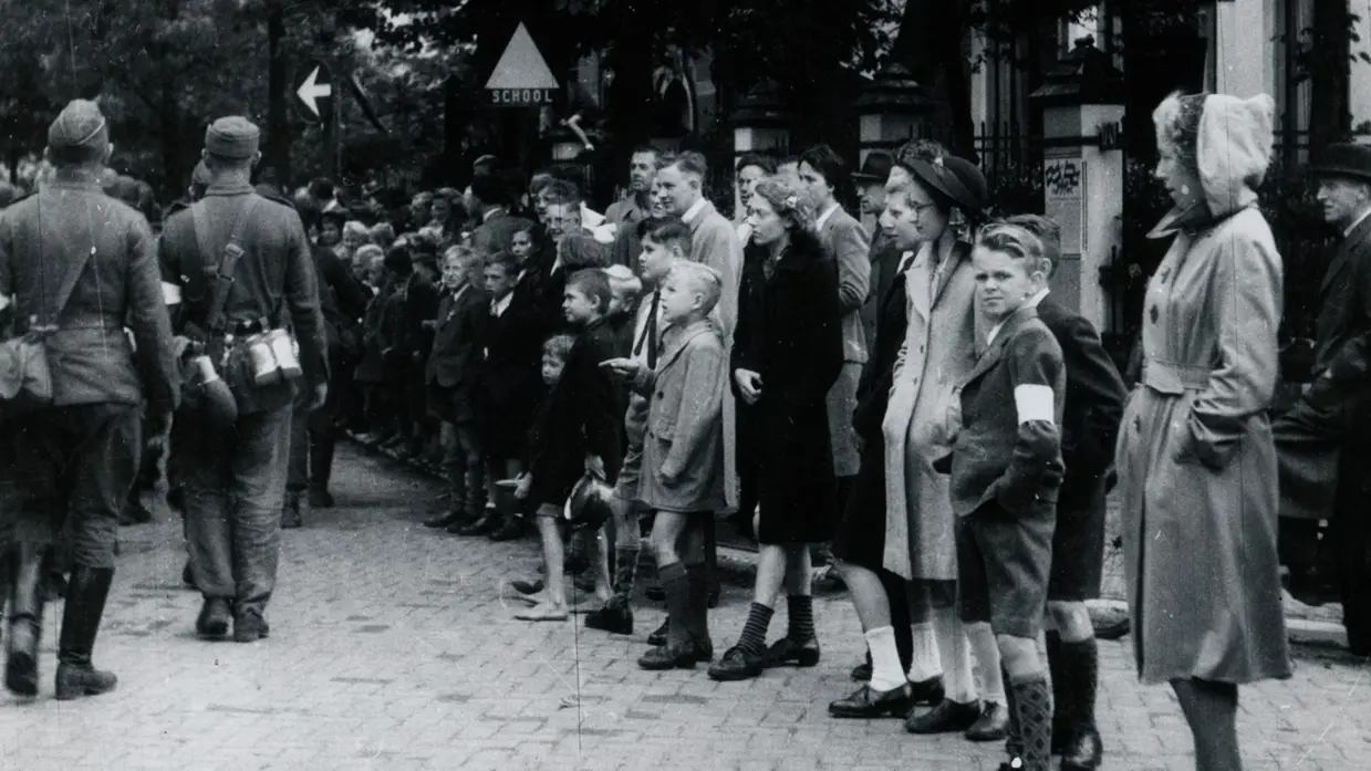 Mensen staan in een rij langs een straat, met soldaten en een bord ‘school’ op de achtergrond.