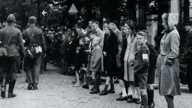 Mensen staan in een rij langs een straat, met soldaten en een bord ‘school’ op de achtergrond.