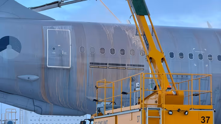 Een gele de-icing truck spuit vanaf hoogte vloeistof op de vleugel van een groot vliegtuig op een besneeuwde luchthaven.