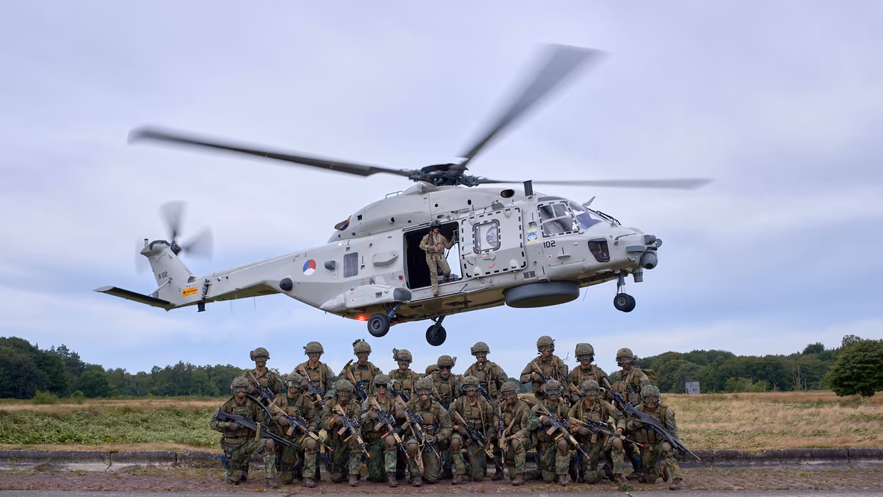Een groep mariniers poseert voor een NH90 die achter hen in de lucht hangt.