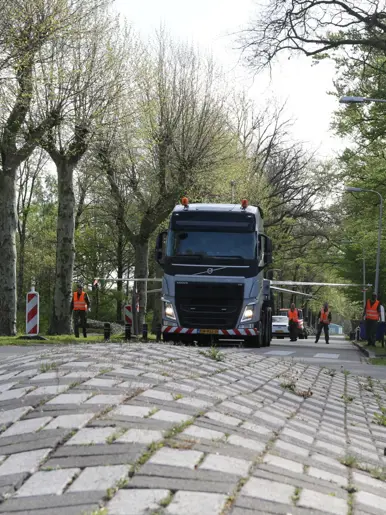 Een grote vrachtwagen rijdt over een weg met tegels, omringd door bomen en verkeersborden, met arbeiders in oranje vesten. 