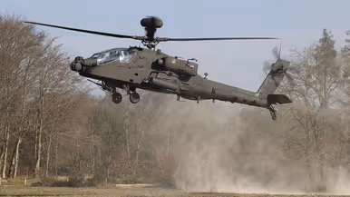 Een zwarte gevechtshelikopter met zware bewapening en draaiende rotorbladen staat op een grasveld tussen kale bomen in een bosrijke omgeving. Rechts: Een militaire helikopter vliegt laag boven een open veld, veroorzaakt stofwolken met ingeladen wapens en draaiende rotorbladen tussen kale bomen.