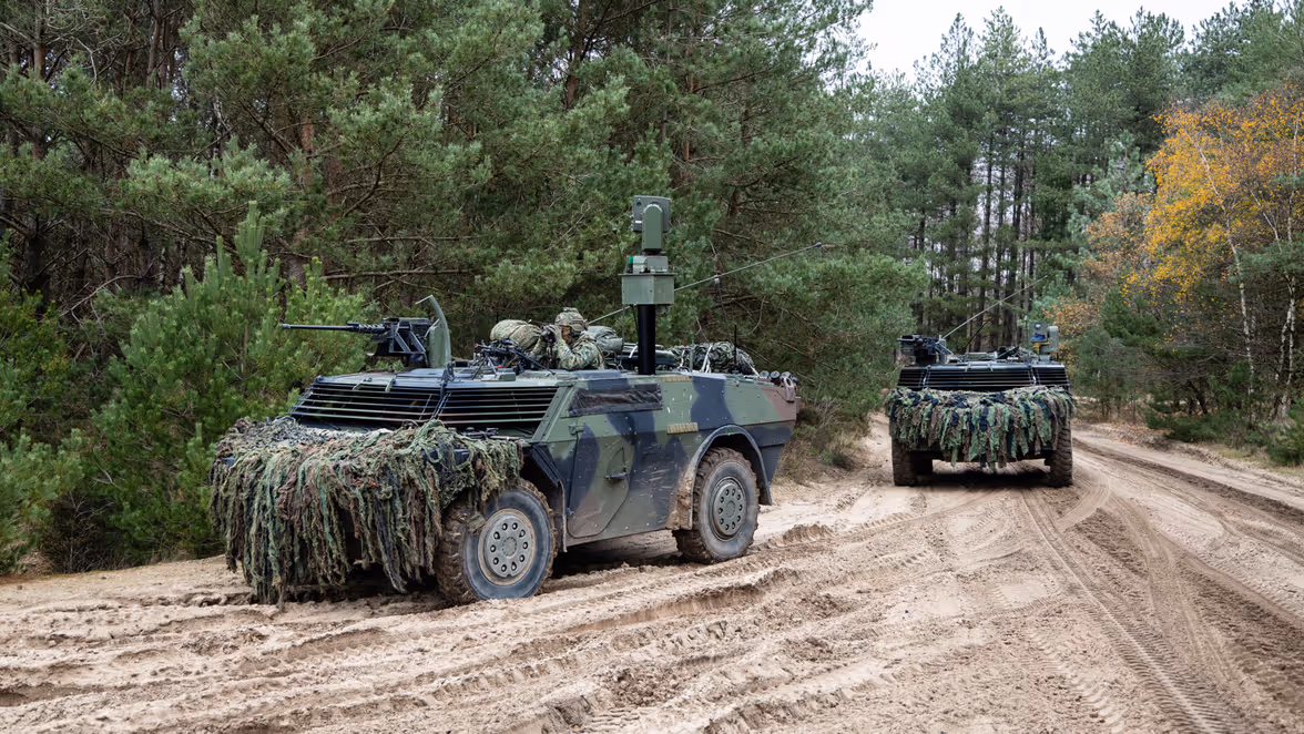 Twee militaire pantservoertuigen, bedekt met camouflagenetten, rijden over een zanderig bospad, omringd door dichte groene bomen en struiken. 
