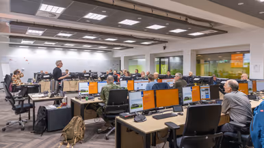  De facilitator van de CDF staat voor de groep en maakt gebruik van zijn handen om de situatie uit te leggen. De aanwezige burgers en militairen luisteren naar hem.