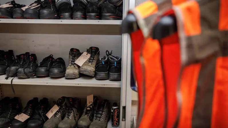  Een grijze kast met daarin veiligheidsschoenen. Daarvoor staat een rek met feloranje veiligheidshesjes.