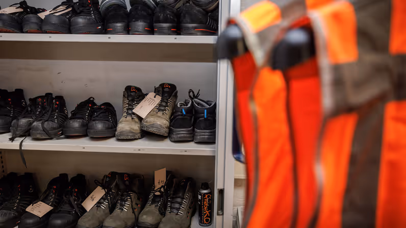  Een grijze kast met daarin veiligheidsschoenen. Daarvoor staat een rek met feloranje veiligheidshesjes.