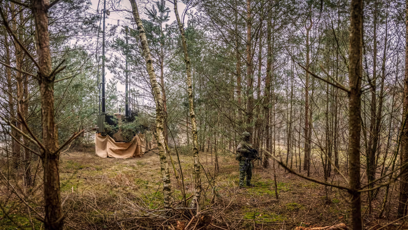 Een militair houdt staand en gewapend de wacht achter een Bushmaster, die verscholen in het groen staat.  