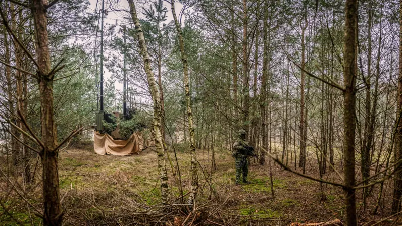 Een militair houdt staand en gewapend de wacht achter een Bushmaster, die verscholen in het groen staat.  