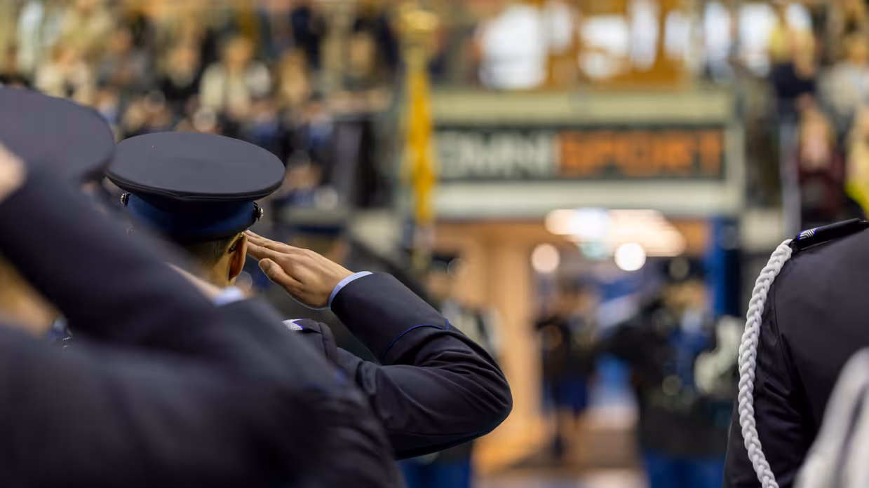 Marechaussees in ceremonieel tenue staan in de houding, met op de achtergrond wazig een bordje met daarop de naam van de sporthal, Omnisport.