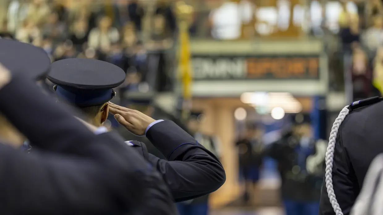 Marechaussees in ceremonieel tenue staan in de houding, met op de achtergrond wazig een bordje met daarop de naam van de sporthal, Omnisport.