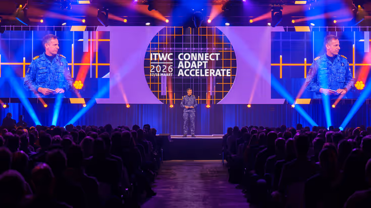 Viceadmiraal Jan Willem Hartman spreekt voor een gevulde zaal. Zijn hoofd is tevens te zien op de twee beeldschermen aan weerszijden van hem.