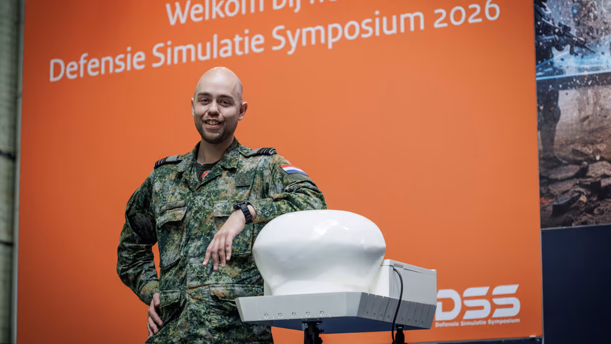 Een militair poseert voor een groot beeldscherm met afbeelding van symposium.