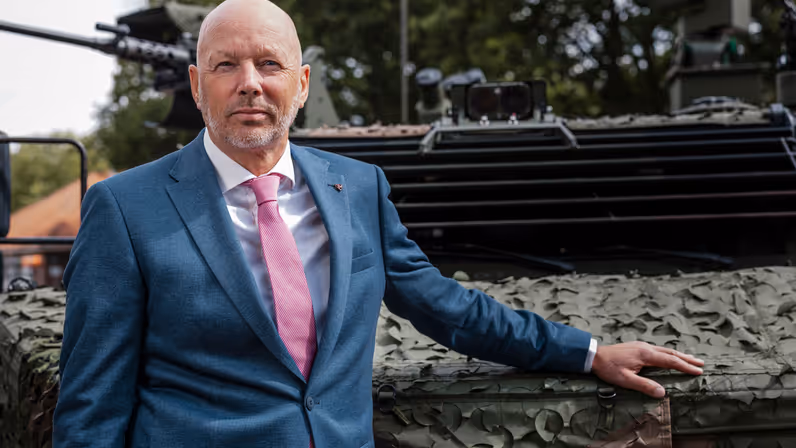 Een kalende man in net pak poseert met zijn hand op een militair voertuig. 