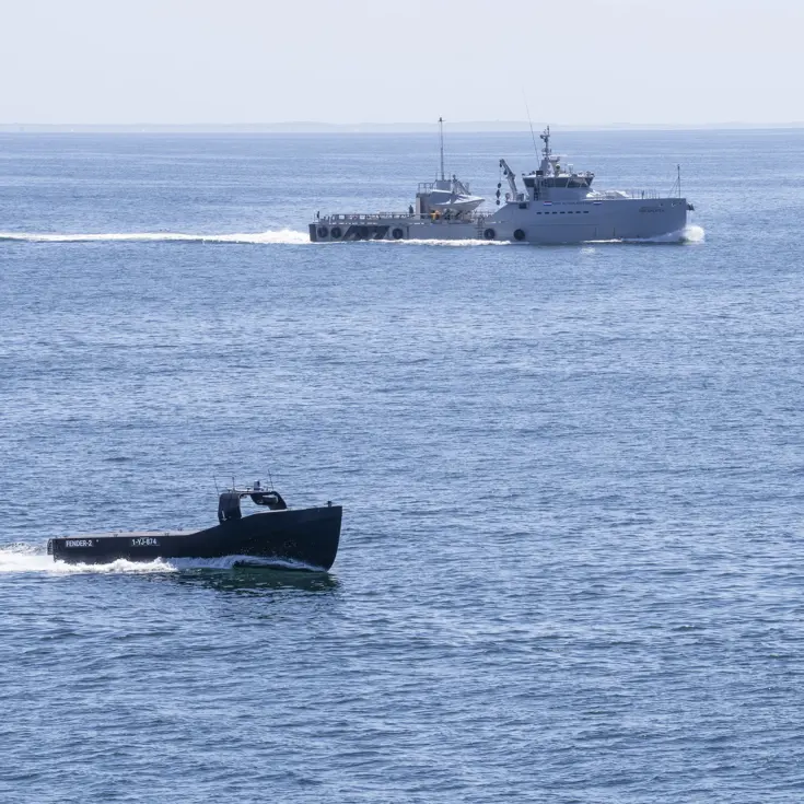 Een onbemand schip vaart op zee met op de achtergrond een patrouillevaartuig.