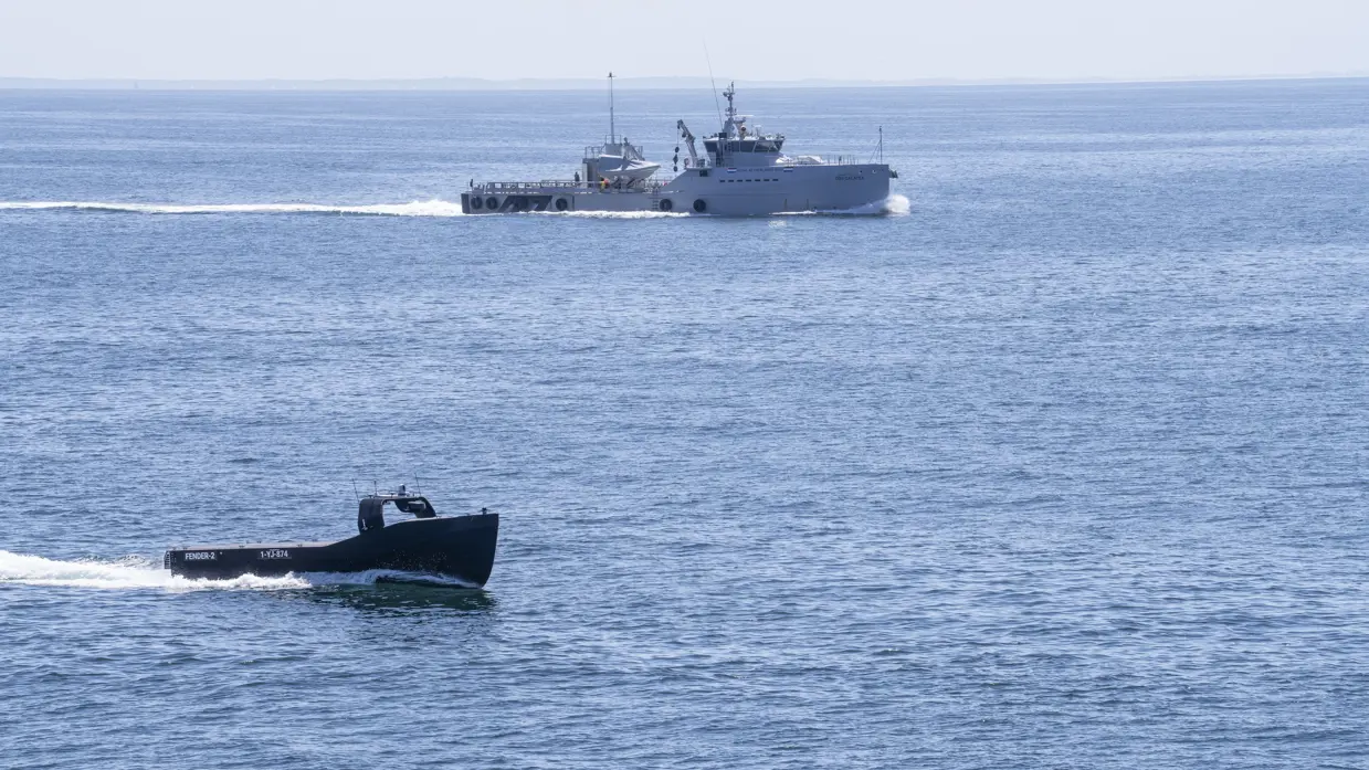Een onbemand schip vaart op zee met op de achtergrond een patrouillevaartuig.
