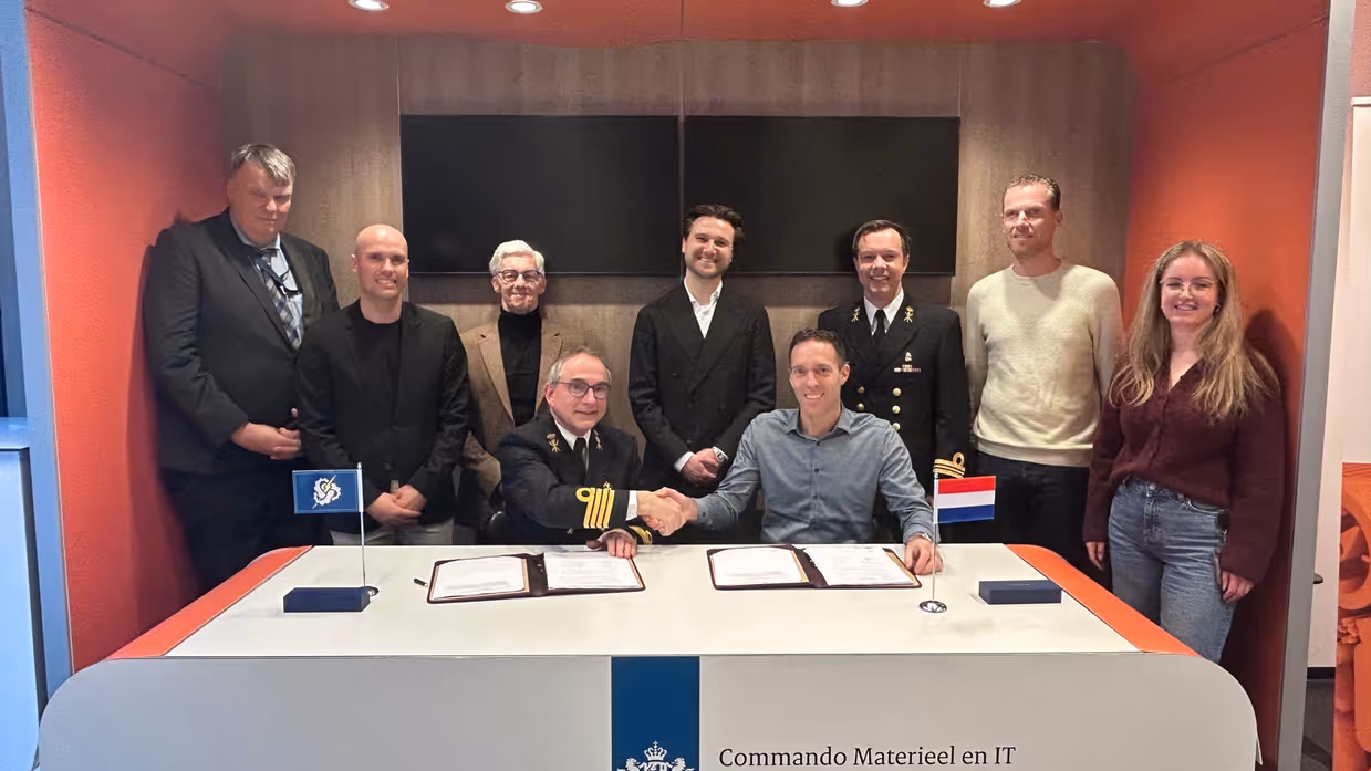  2 heren achter een desk, van wie 1 in militair DT,  schudden elkaar de hand met daarachter 6 mannen en een vrouw.