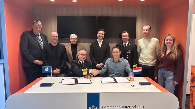  2 heren achter een desk, van wie 1 in militair DT,  schudden elkaar de hand met daarachter 6 mannen en een vrouw.