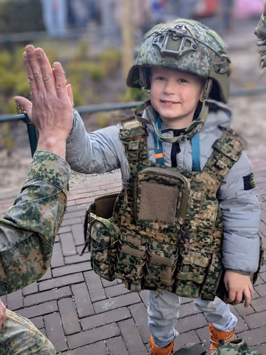 Links: Twee glimlachende kinderen kijken onder een grote, groene camouflagenetten-tent vandaan, die buiten voor huizen is opgebouwd en lijkt op een militaire schuilplaats. Rechts: Een glimlachend kind in een militair kogelwerend vest en helm geeft een volwassene in uniform een high-five op een buitenlocatie.