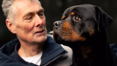 Links een man in uniform. Hij omarmt een grote zwart-bruine hond met opgetrokken oren. Achtergrond toont een bosrijke omgeving met een metalen hek. Rechts een glimlachende man in uniform staat naast een zwarte Rottweiler met open bek. Bosrijke omgeving op de achtergrond, met een ontspannen en vriendelijke sfeer. 