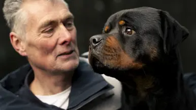 Links een man in uniform. Hij omarmt een grote zwart-bruine hond met opgetrokken oren. Achtergrond toont een bosrijke omgeving met een metalen hek. Rechts een glimlachende man in uniform staat naast een zwarte Rottweiler met open bek. Bosrijke omgeving op de achtergrond, met een ontspannen en vriendelijke sfeer. 