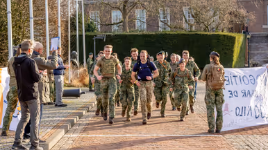 Links een groep mensen in militaire kleding loopt rennend over een keienpad, omringd door bomen en een gebouw. Aan de zijkant houden twee mannen een spandoek vast. Het is een zonnige dag. Rechts: twee mannen omarmen elkaar hartelijk buiten in de zon. De ene draagt een groene jas, de andere een donkerblauw sportshirt met een oranje logo. Op de achtergrond staan twee oudere mannen. 