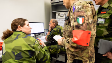 Links: Militairen in uniform werken in een kantoorruimte met laptops en papieren. Een man met Nederlandse vlag op zijn arm staat te luisteren. Kaarten hangen aan de muur. Rechts: Militairen in verschillende uniformen bespreken informatie achter een computer. Een vrouw met Italiaanse vlag op haar mouw kijkt toe, terwijl een man nadenkt.