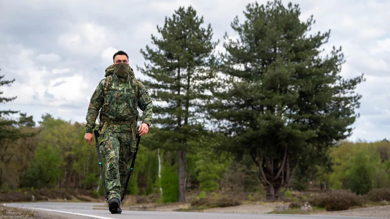 Een militair in camouflage met rugzak en stokken loopt over een weg met dennenbomen en een bewolkte lucht. 