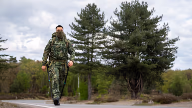 Een militair in camouflage met rugzak en stokken loopt over een weg met dennenbomen en een bewolkte lucht. 
