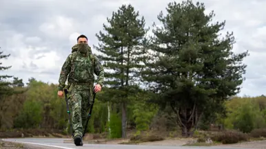 Een militair in camouflage met rugzak en stokken loopt over een weg met dennenbomen en een bewolkte lucht. 