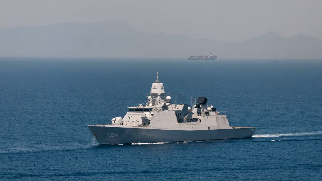 Een marineschip vaart op zee met in de verte een ander schip en hoge bergen.