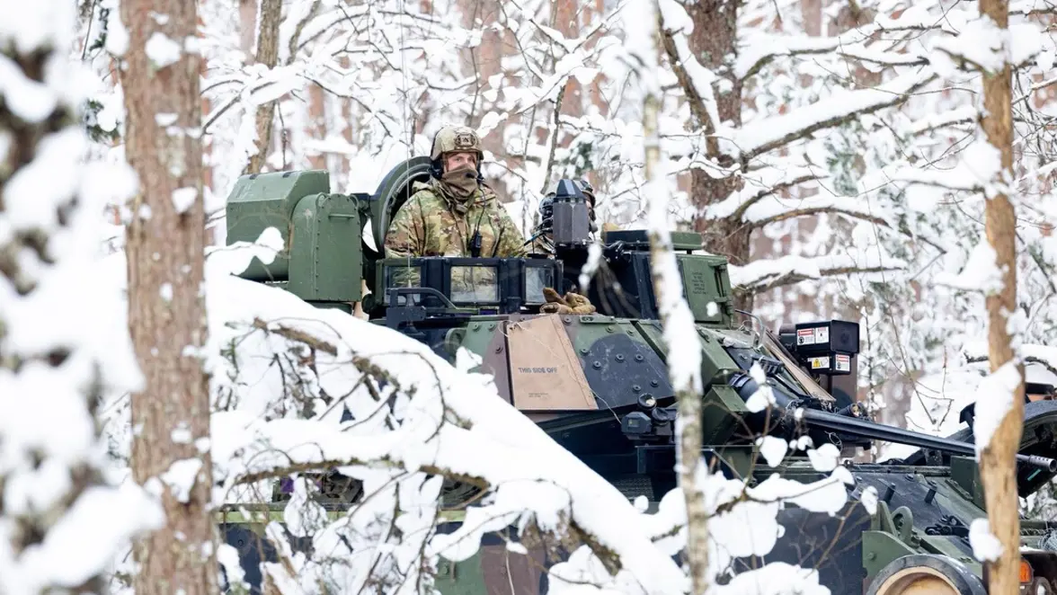 Een soldaat in winterkleding zit bovenop een gepantserd militair voertuig tussen besneeuwde bomen, omringd door sneeuw en rustige, witte natuur. 
