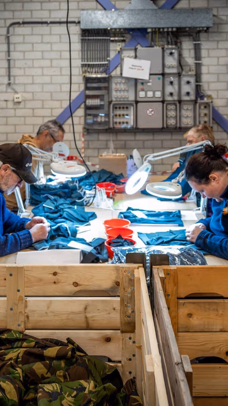 Vier personen, van wie een met pet, werken met kleding aan een tafel.  