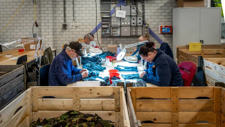 Vier personen, van wie een met pet, werken met kleding aan een tafel.  