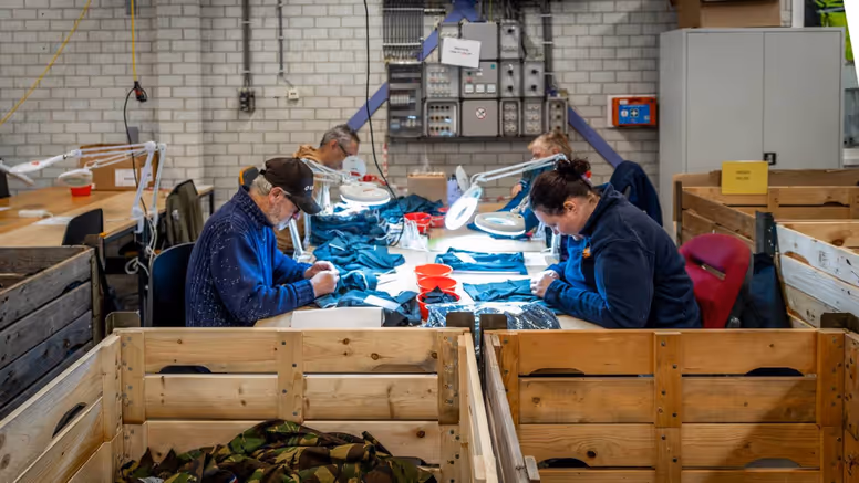 Vier personen, van wie een met pet, werken met kleding aan een tafel.  