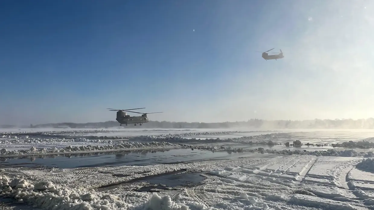 Twee helikopters vliegen laag over een besneeuwde, modderige en ijsbedekte landingsbaan onder een heldere, blauwe hemel met zonnig licht. 