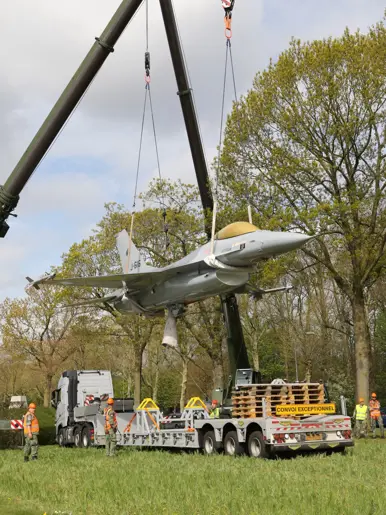 Een grote kraan tilt een straaljager op boven een vrachtwagen, omringd door bomen en arbeiders in veiligheidskleding. 