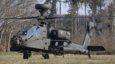 Een zwarte gevechtshelikopter met zware bewapening en draaiende rotorbladen staat op een grasveld tussen kale bomen in een bosrijke omgeving. Rechts: Een militaire helikopter vliegt laag boven een open veld, veroorzaakt stofwolken met ingeladen wapens en draaiende rotorbladen tussen kale bomen.