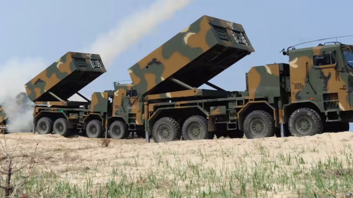 Een groot militair raketwerpervoertuig met camouflagekleuren schiet raketten af, met rook en vuur zichtbaar, op een zanderige heuvel met groen gras. 