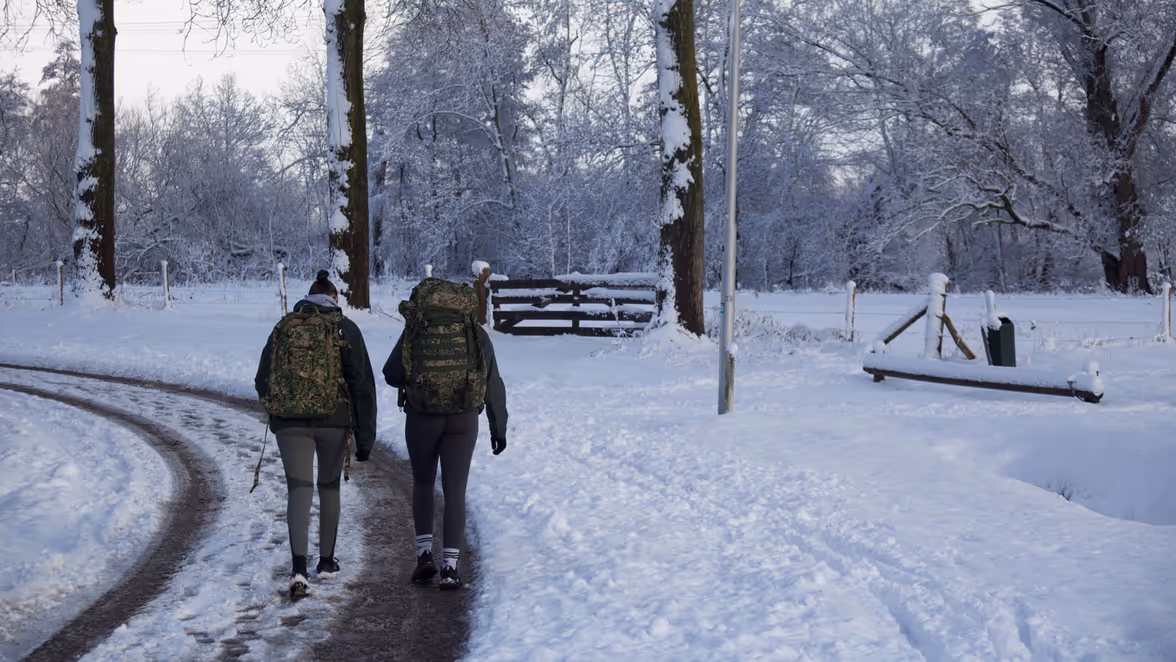 Twee mensen lopen met rugzakken in militaire print over een besneeuwd, rustig pad in een winterbos, omringd door sneeuwbedekte bomen en een bankje. 