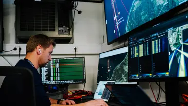 Een man werkt in een controlekamer met schermen vol kaarten, radarbeelden en gegevens, omringd door apparatuur.