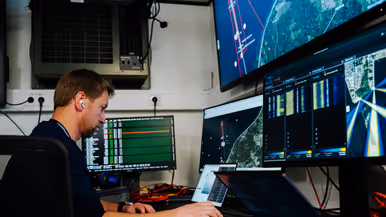 Een man werkt in een controlekamer met schermen vol kaarten, radarbeelden en gegevens, omringd door apparatuur.