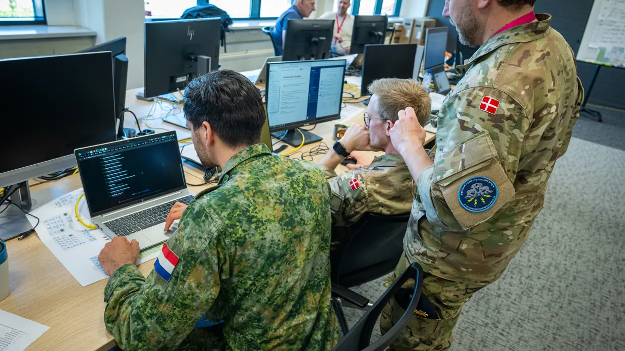 Twee Deense militairen, van wie een staand met een Nederlandse collega naar twee beeldschermen kijkt. 